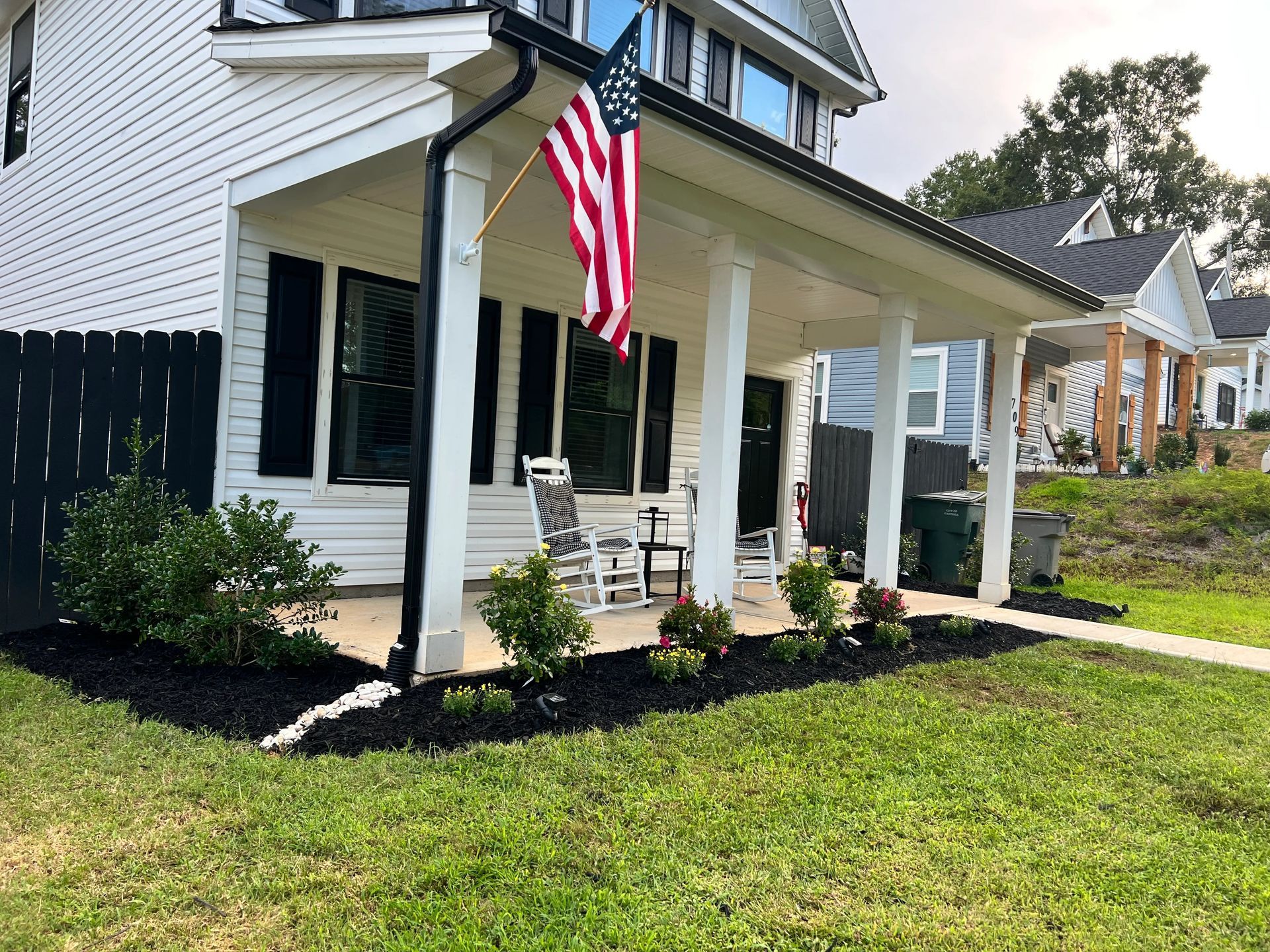 White two-story house with a front porch, American flag, rocking chairs, and landscaped flower beds with black mulch.