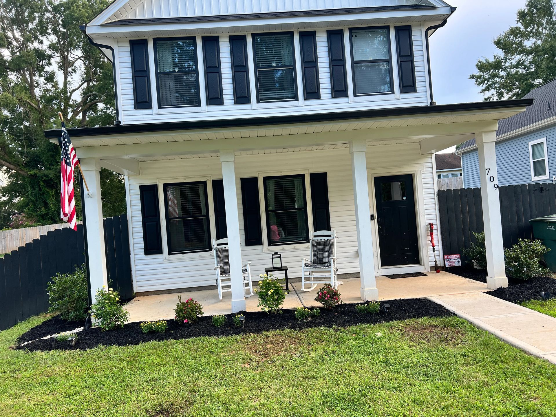 A two-story white house with black shutters, a full-width front porch, two rocking chairs, and an American flag.