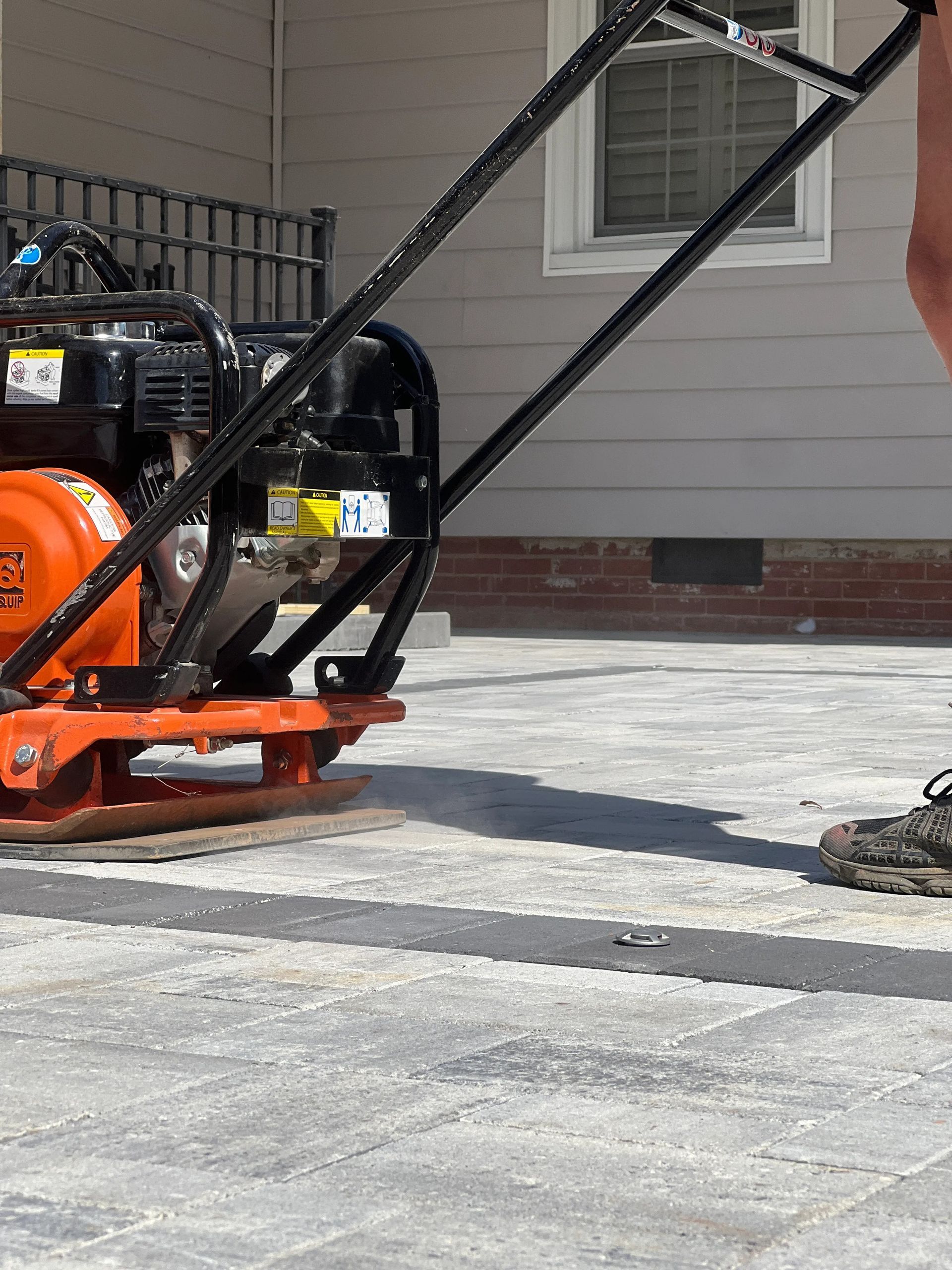A person uses a walk-behind orange plate compactor to smooth newly installed pavers on a residential patio.