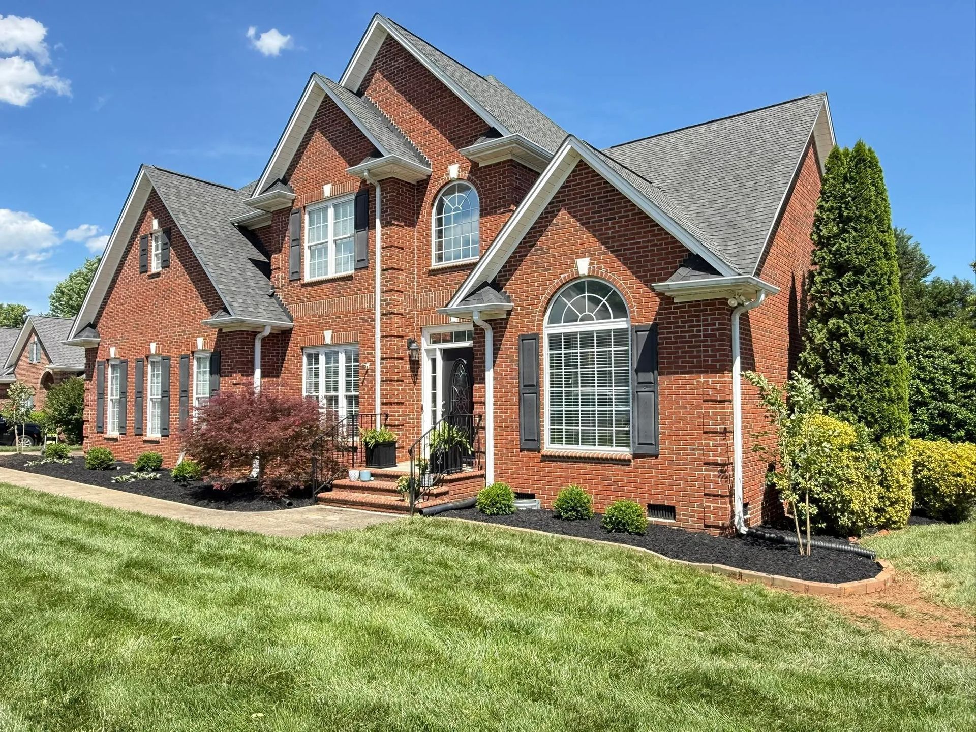A two-story red brick house with a gray shingled roof, arched windows, and neatly landscaped lawn under a blue sky.