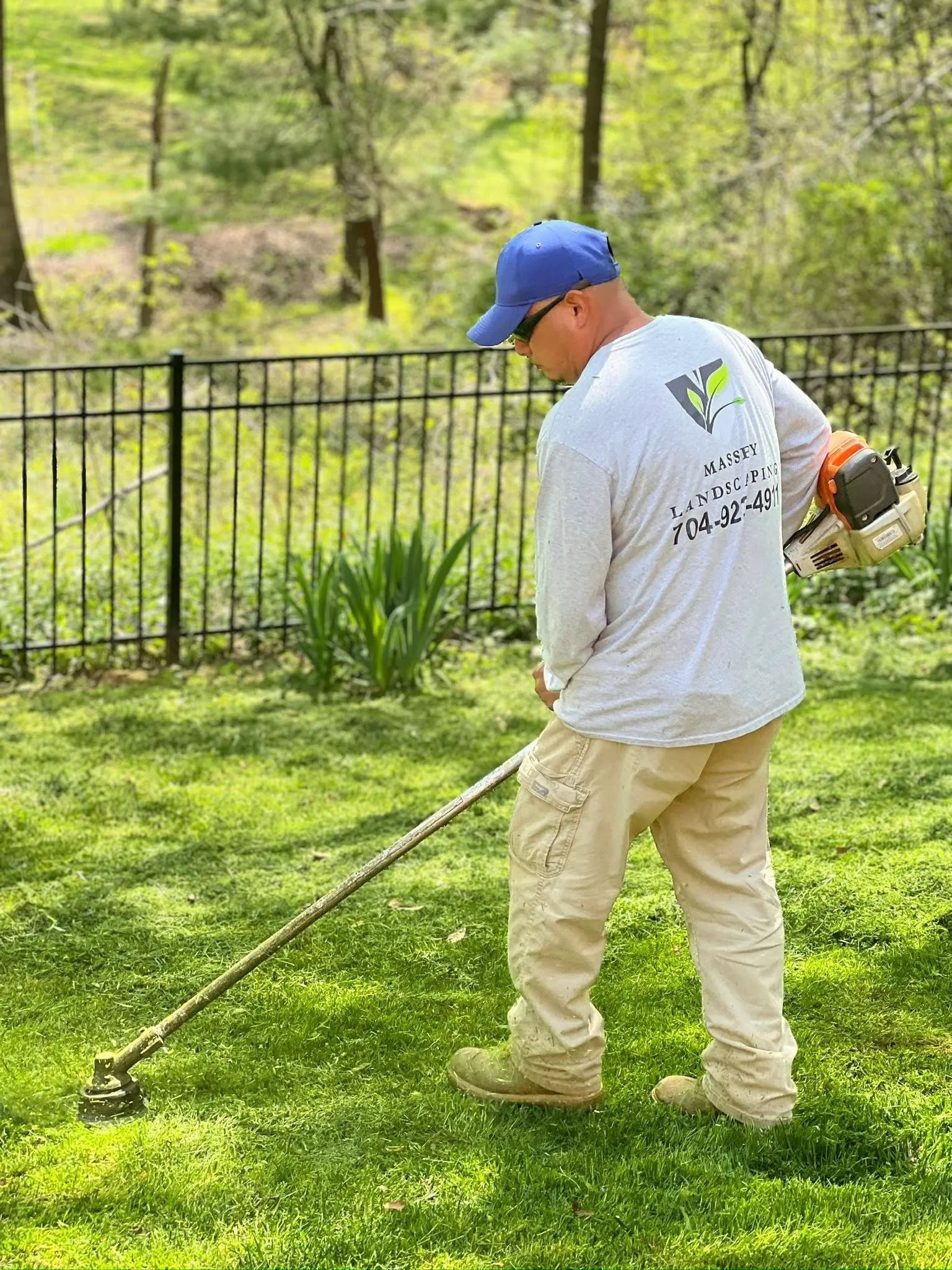 A person wearing a blue cap and light-colored clothing uses a string trimmer to clear grass in a grassy yard.