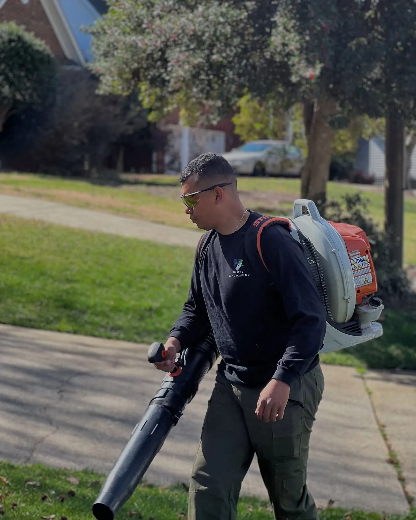 A person wearing safety glasses and a backpack leaf blower walks across a residential lawn on a sunny day.