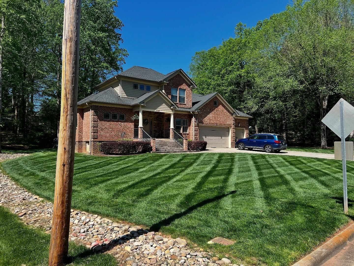 A large, two-story brick house with a manicured, striped lawn and a blue car parked in the driveway on a sunny day.