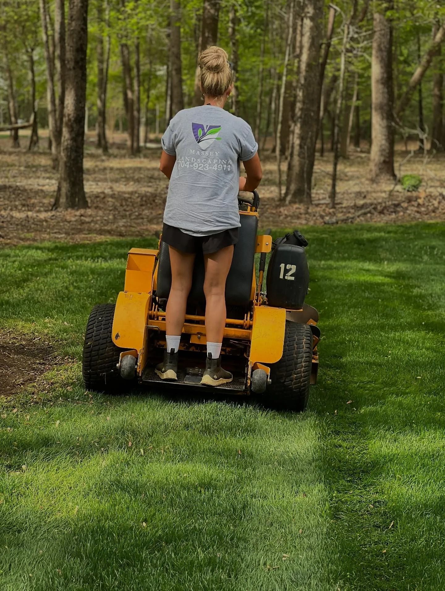 A person stands on a yellow stand-on mower, cutting a strip of grass in a wooded yard.