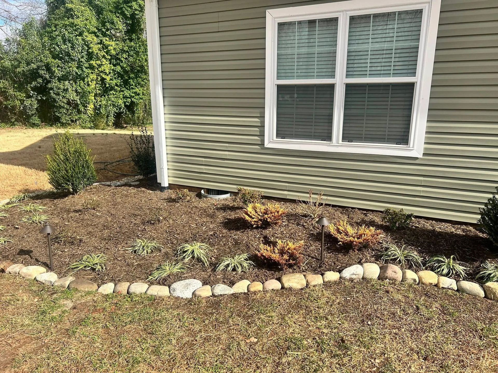 A tan house exterior with a window above a flower bed mulched with brown wood chips and outlined by river stones.