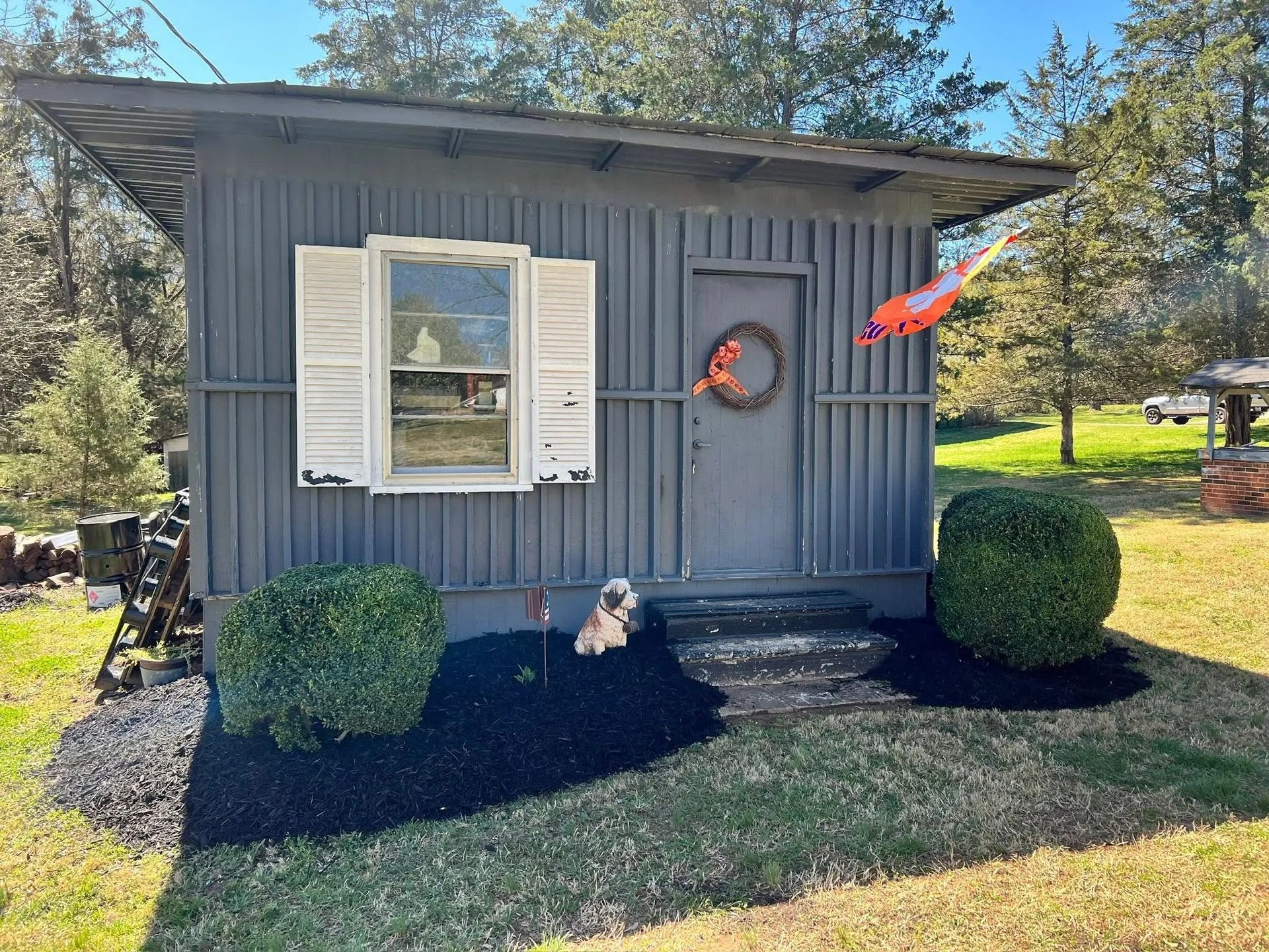 A small, grey, vertical-sided shed with a single window with white shutters, a door with a wreath, and two round bushes.