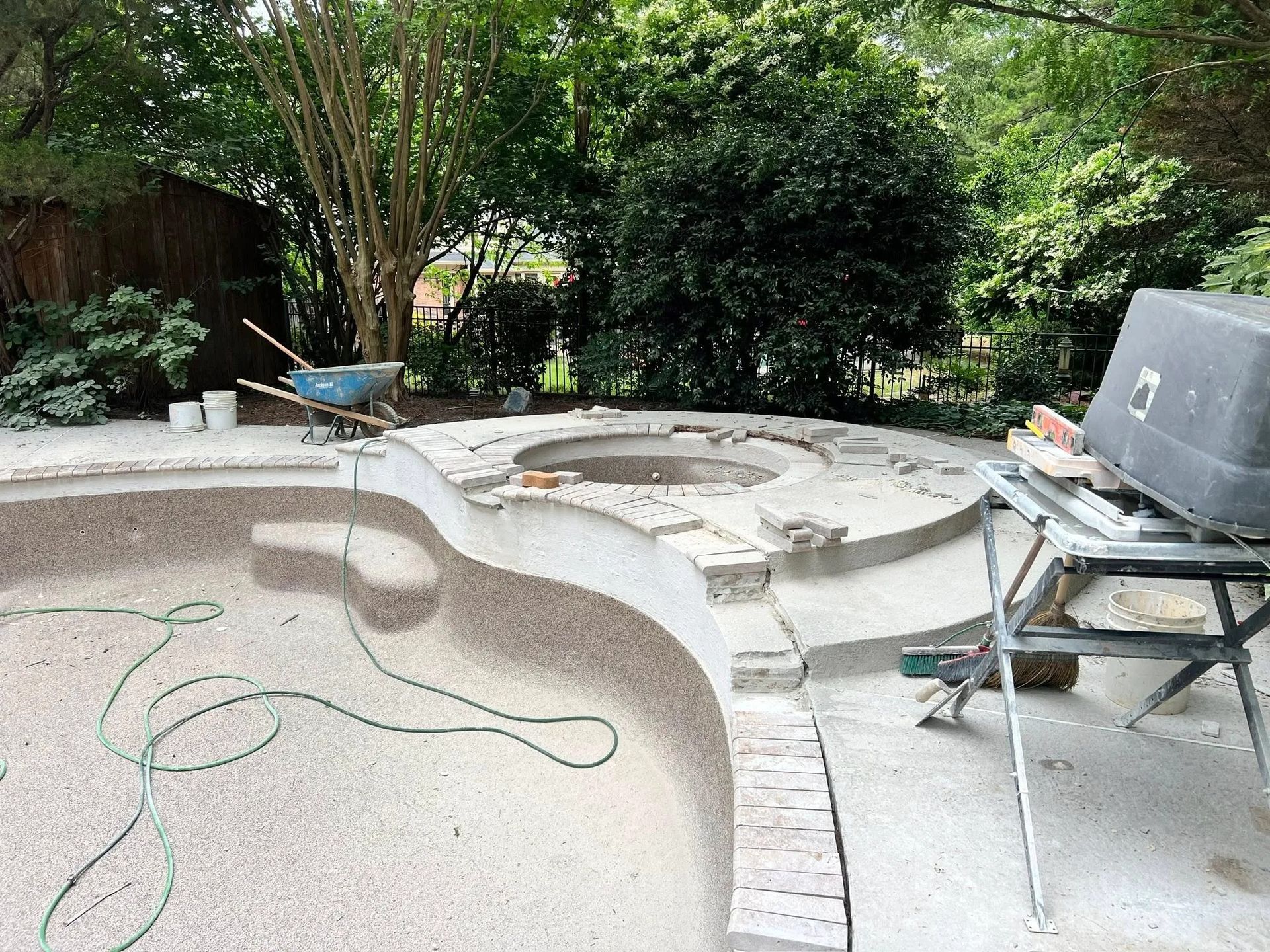 A backyard swimming pool under construction, featuring a partially tiled spa, a wet saw, and a wheelbarrow nearby.