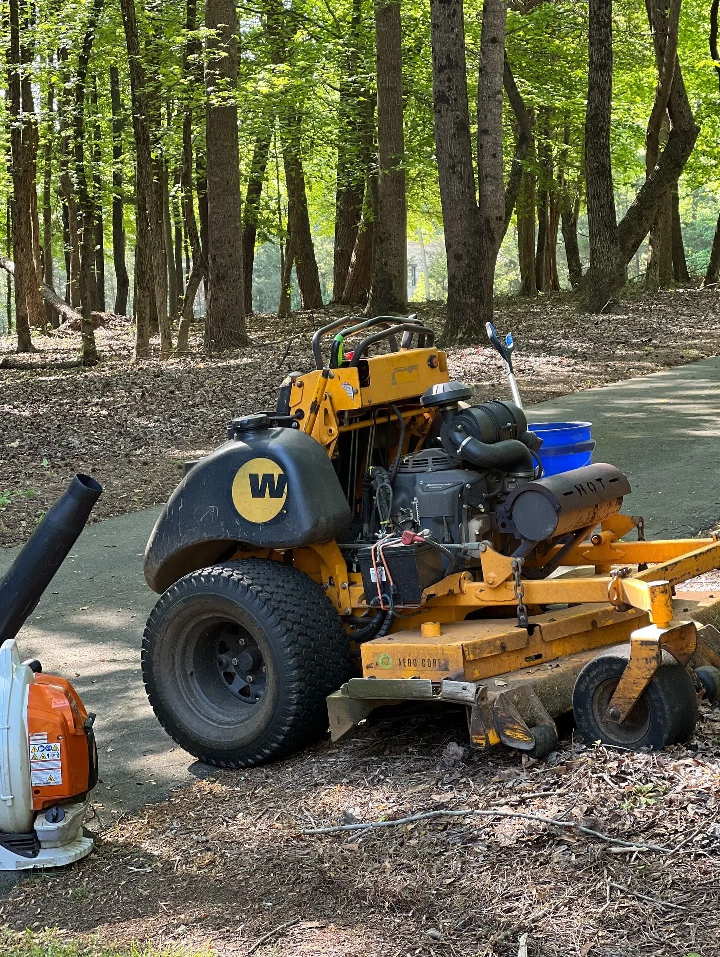 A yellow Wright stand-on lawn mower parked on a path in a wooded area, next to a backpack leaf blower.