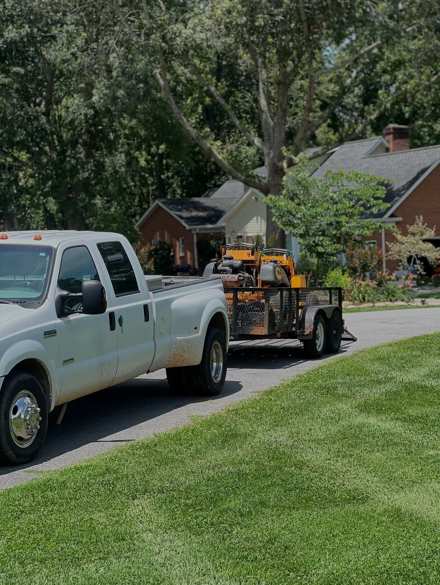 A white pickup truck with a trailer carrying yellow construction equipment parked on a residential street.
