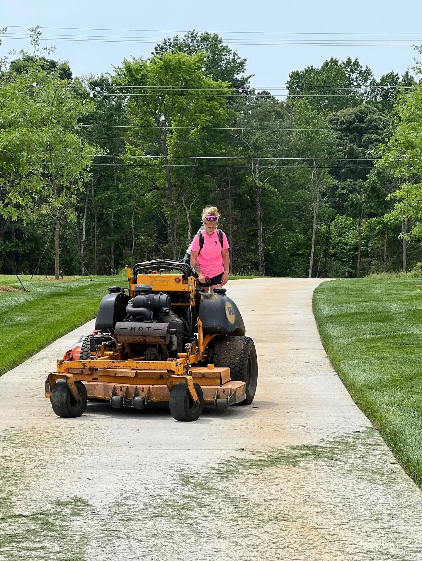 A person in a bright pink shirt rides a yellow commercial zero-turn mower down a gravel driveway lined with green grass.