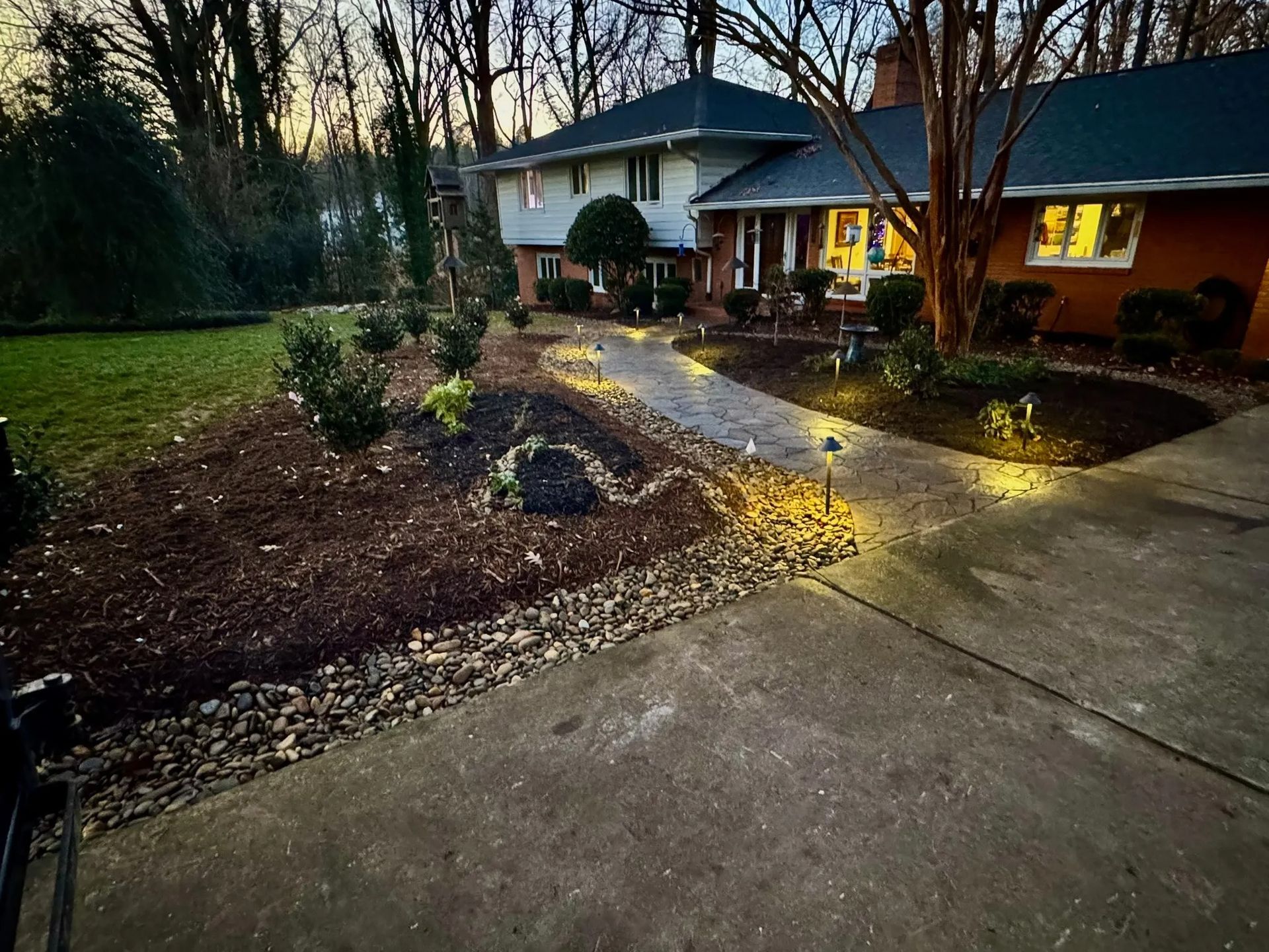 A house at dusk with a stone walkway lined by small lights leading to the front door, bordered by mulch and greenery.