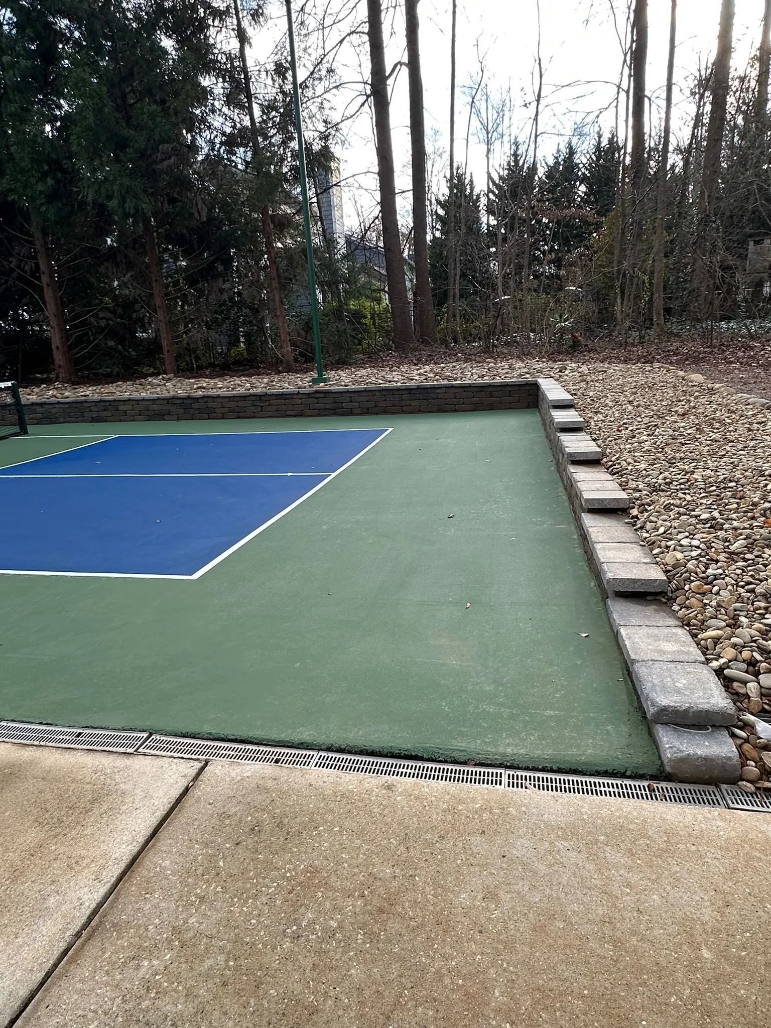 A pickleball court with a blue playing area, green surface, and a stepped retaining wall bordering a wooded area.