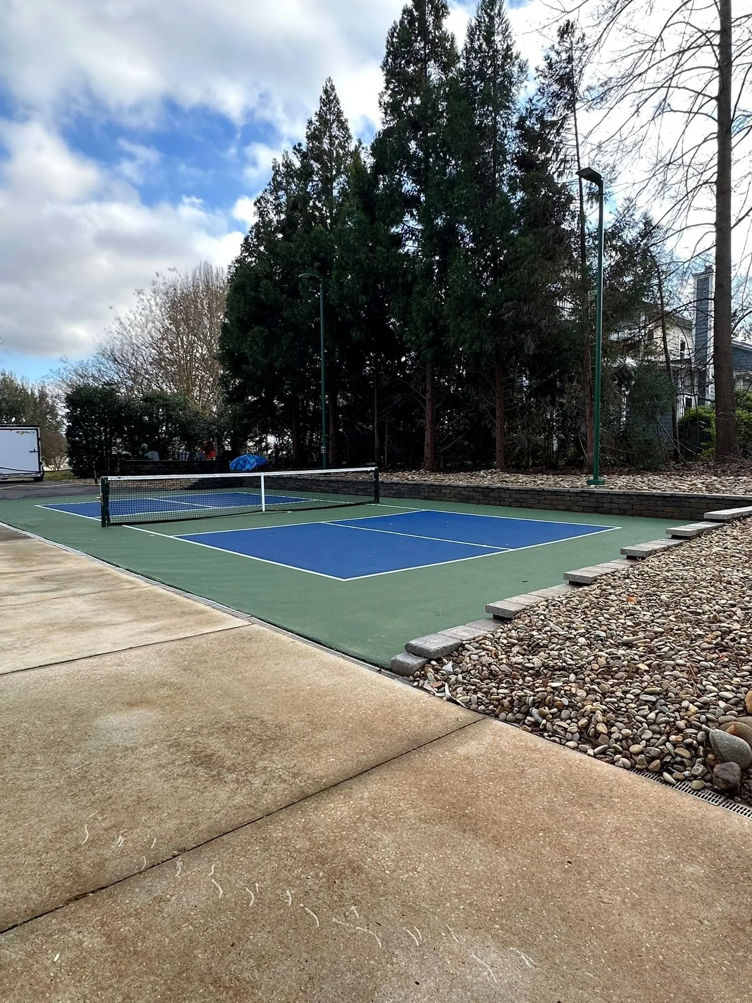 Outdoor pickleball court with a blue playing area, green border, and net, surrounded by trees and gravel.