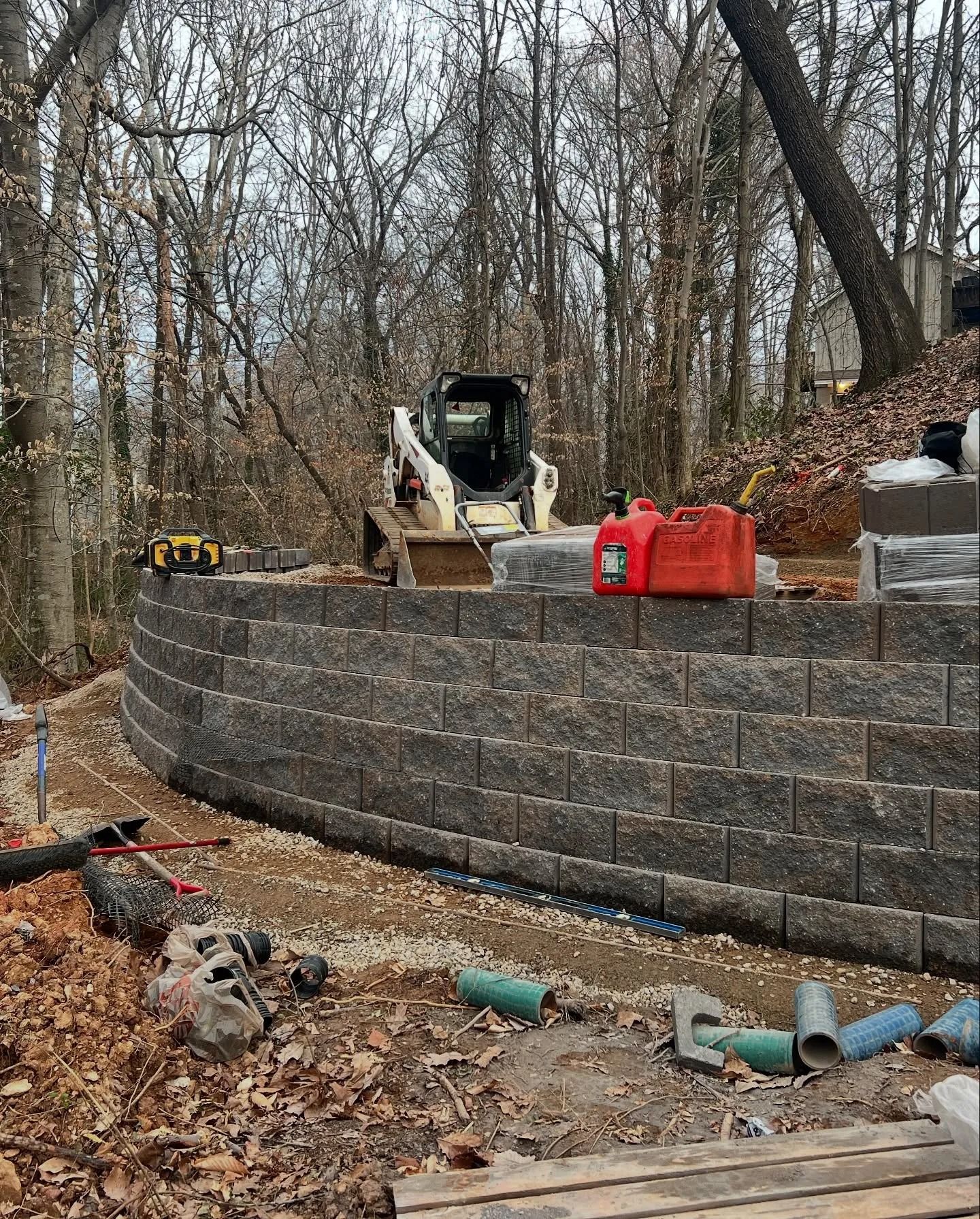 A retaining wall under construction in a wooded area with a skid steer loader, red fuel cans, and drainage pipes nearby.