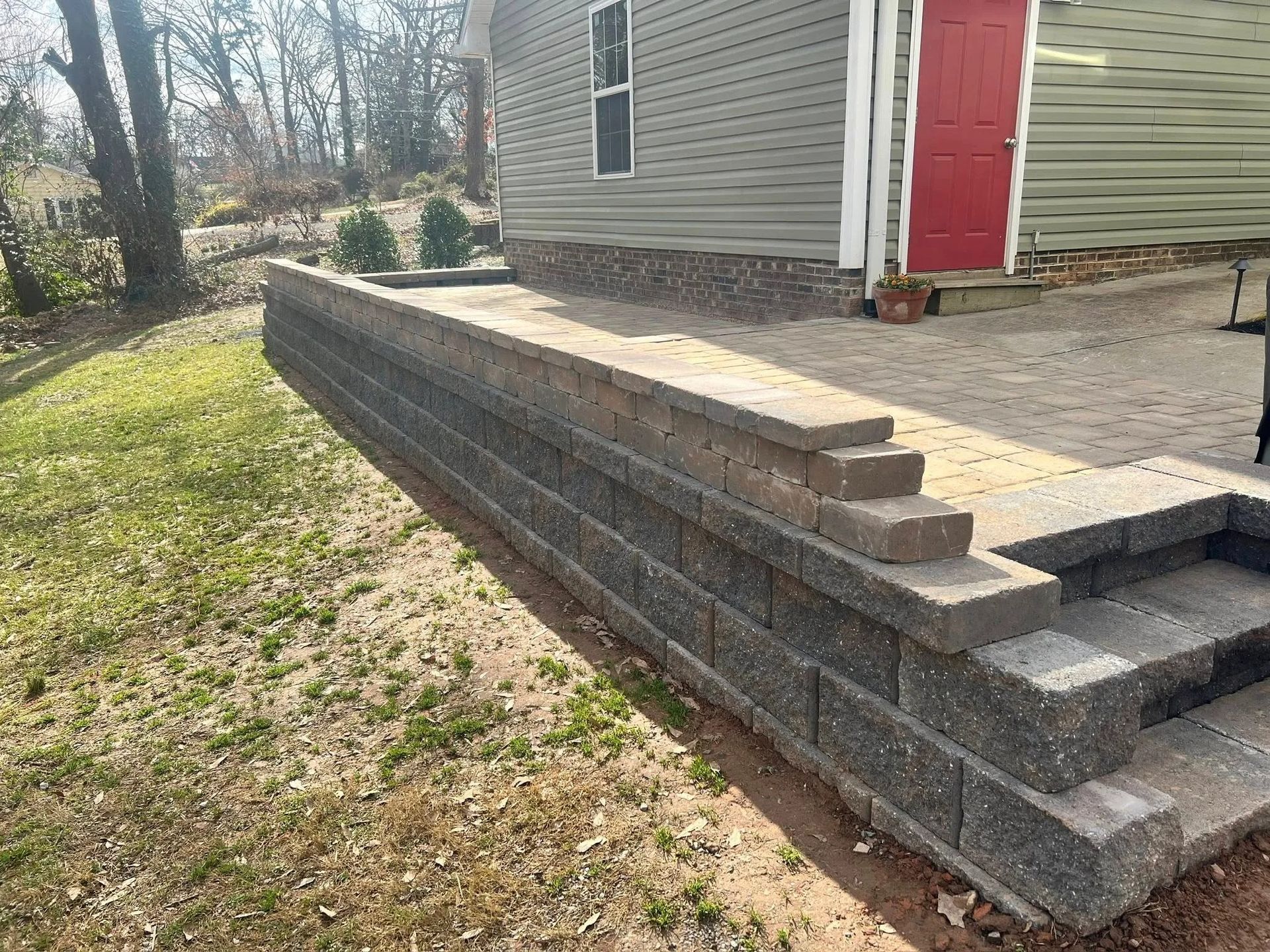 A stone retaining wall with steps leads up to a paved patio next to a house with grey siding and a red door.