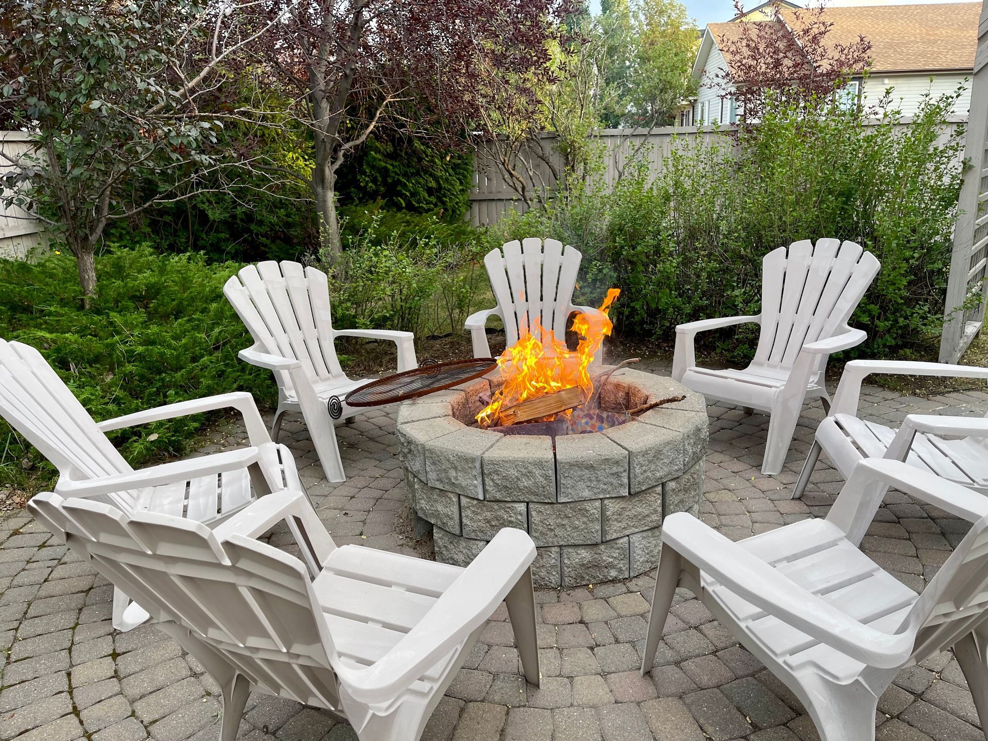 White Adirondack chairs arranged in a circle around a stone fire pit with a glowing fire in a backyard garden setting.