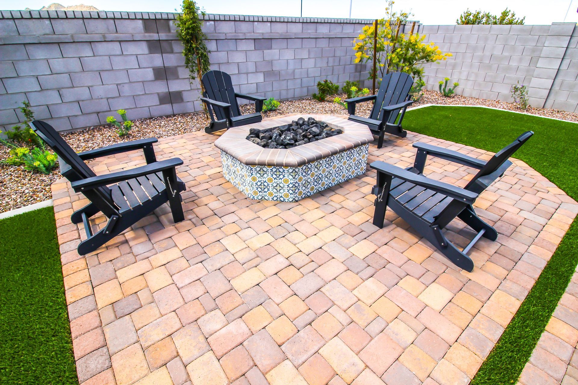 Four black Adirondack chairs surround a tiled, octagonal fire pit on a brick patio in a backyard with artificial turf.
