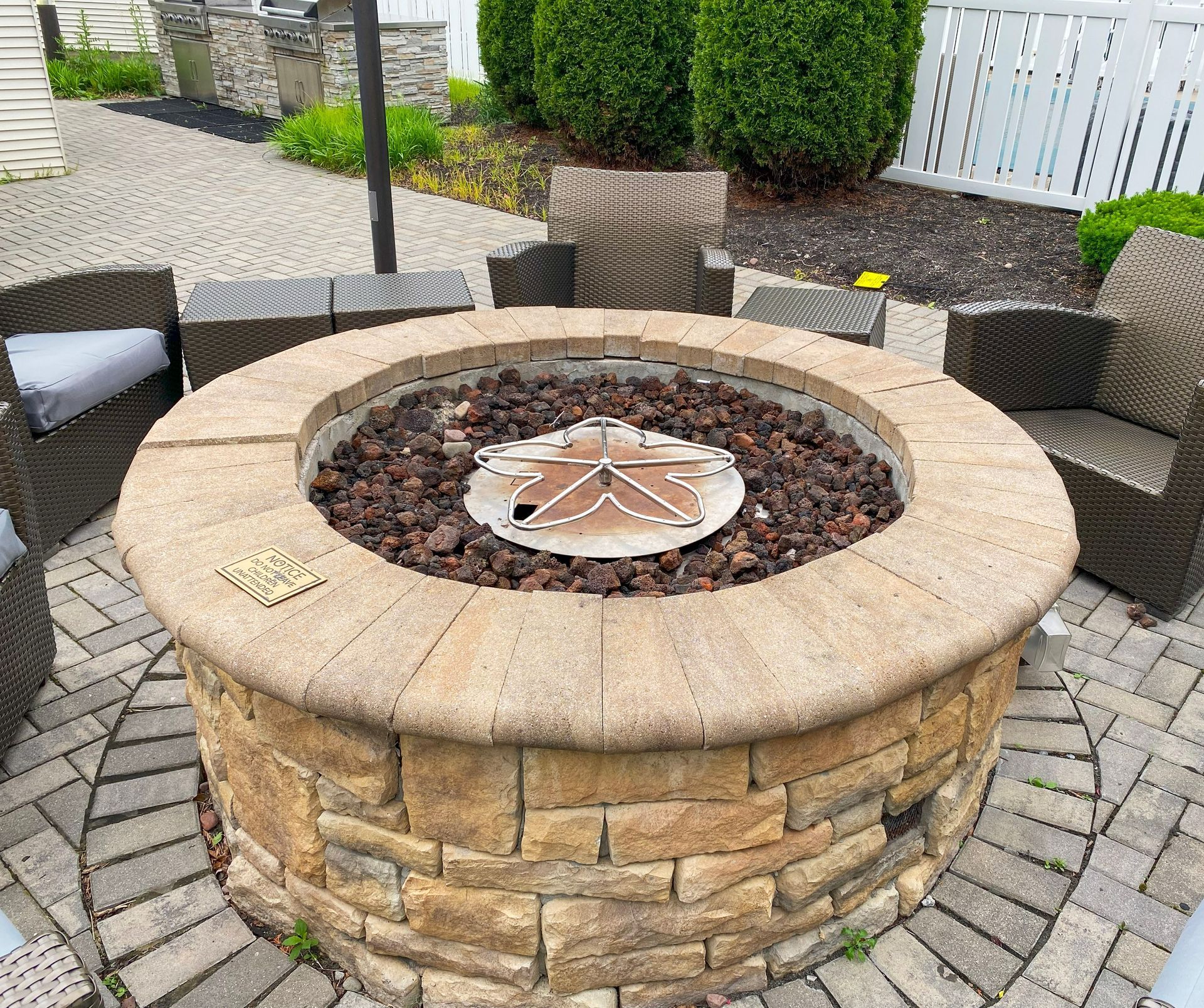 A round stone fire pit with dark lava rocks and a metal star-shaped grate, centered on a brick patio with wicker chairs.
