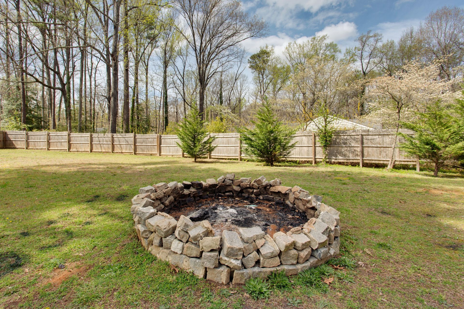 A stone fire pit centered in a grassy backyard, with a wooden fence and tall trees in the background.