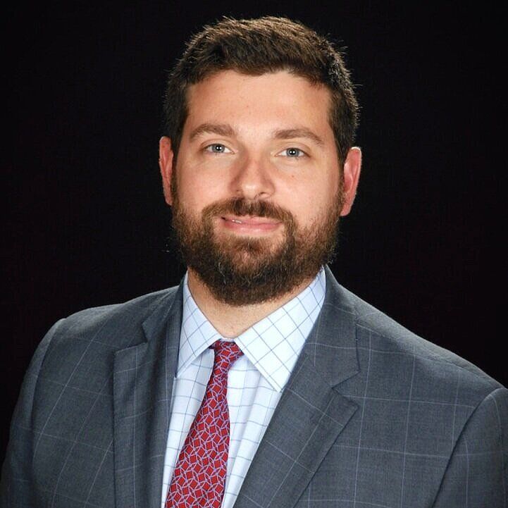 Man in a gray suit and patterned tie, smiling with a full beard, against a dark background.