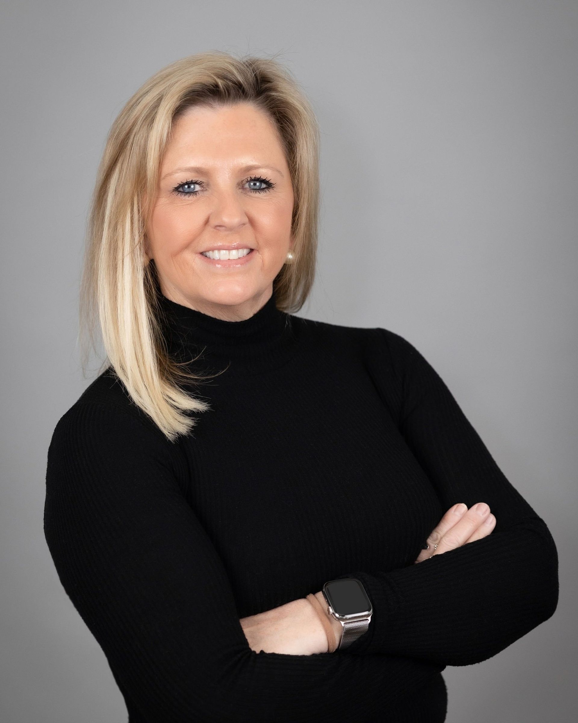 A person with blonde hair wearing a black turtleneck stands with arms crossed against a plain grey background.
