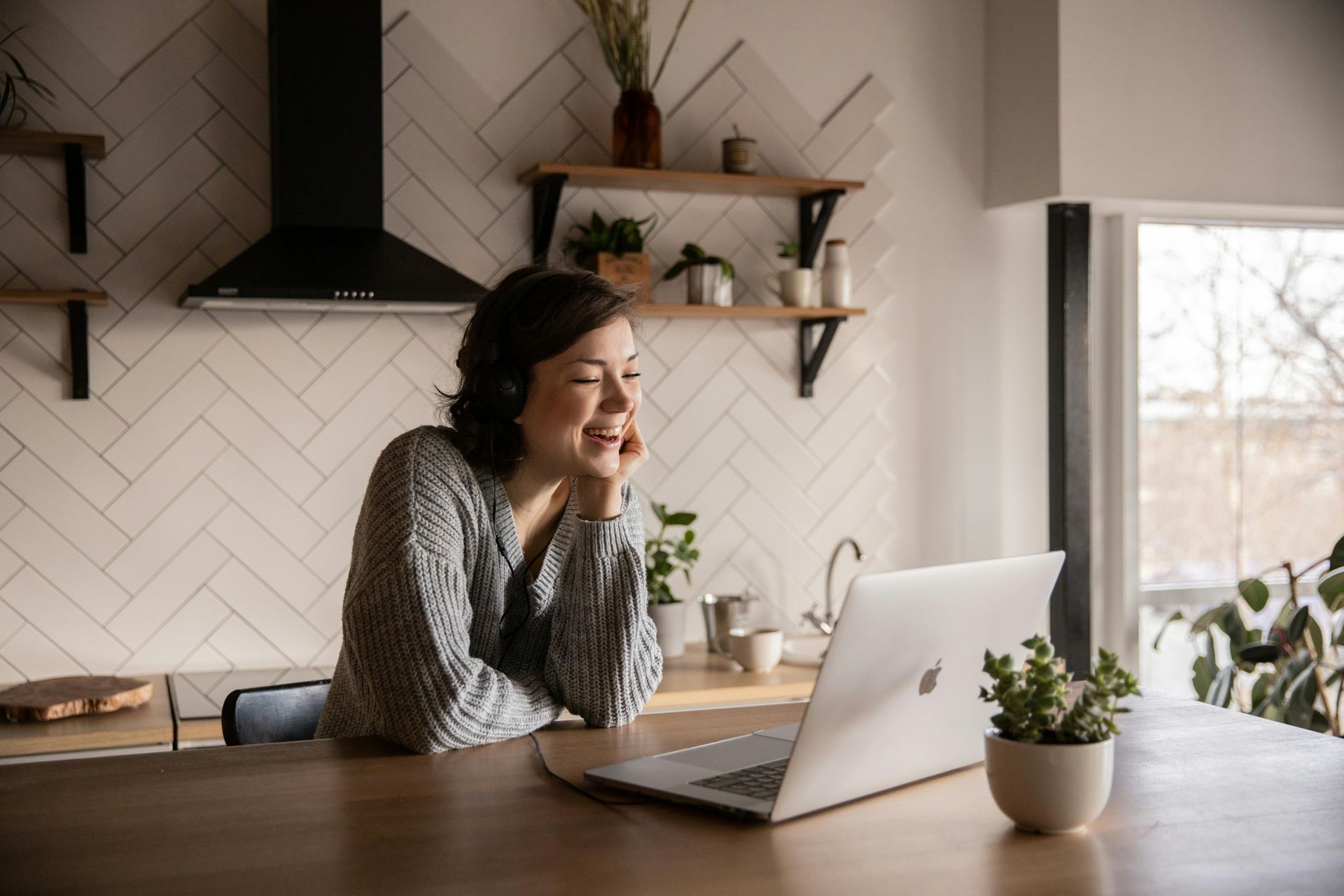 A smiling person sits at a wooden table in a bright kitchen, looking at an open laptop.