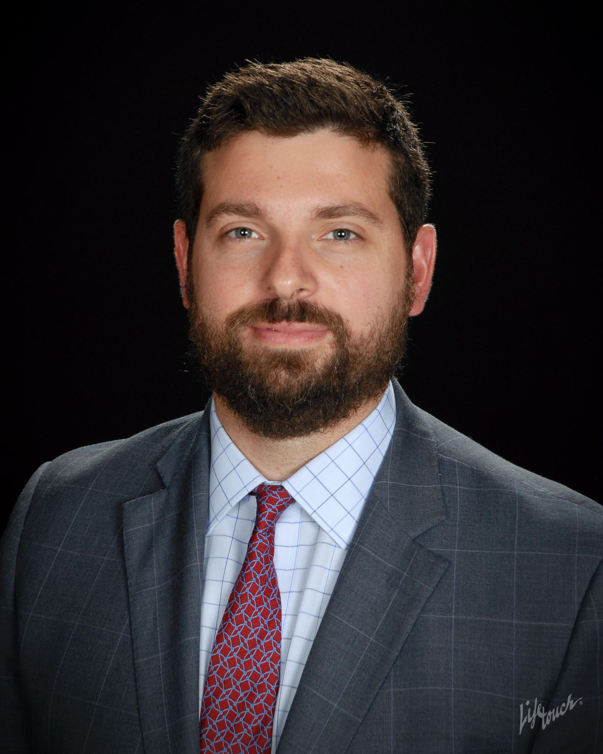 A headshot of a person with a beard, wearing a gray plaid blazer, a light blue checkered shirt, and a red patterned tie.