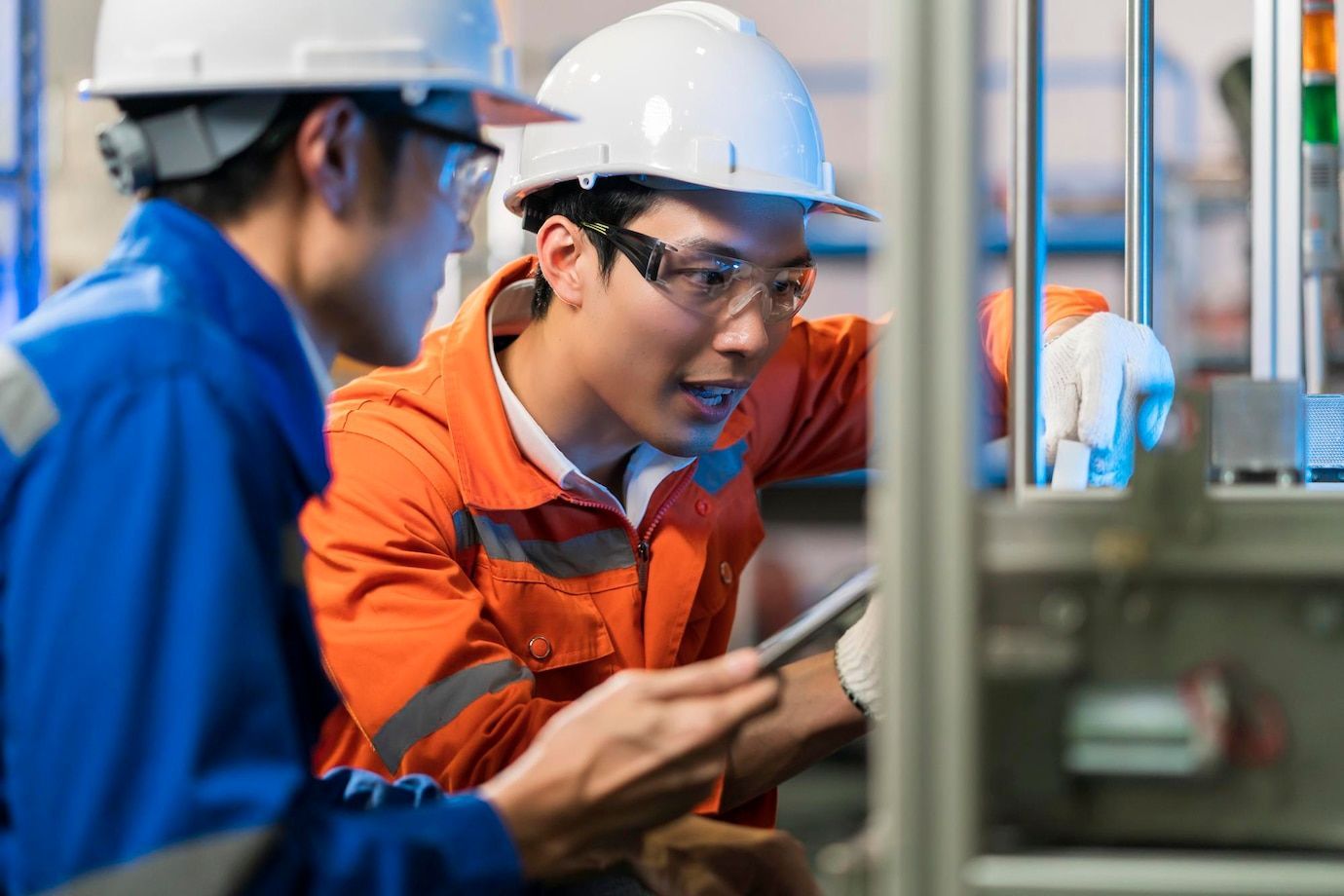 Two men wearing hard hats and safety glasses are working on a machine in a factory.