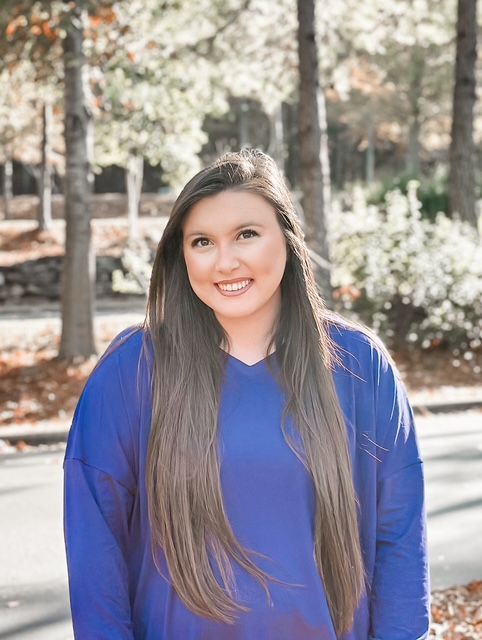 Woman with long brown hair smiles, wearing a blue sweater outside with trees in the background.