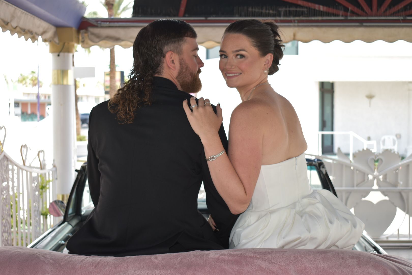 Bride and groom in a convertible. She smiles, wearing a white strapless dress, while he has a mullet hairstyle.