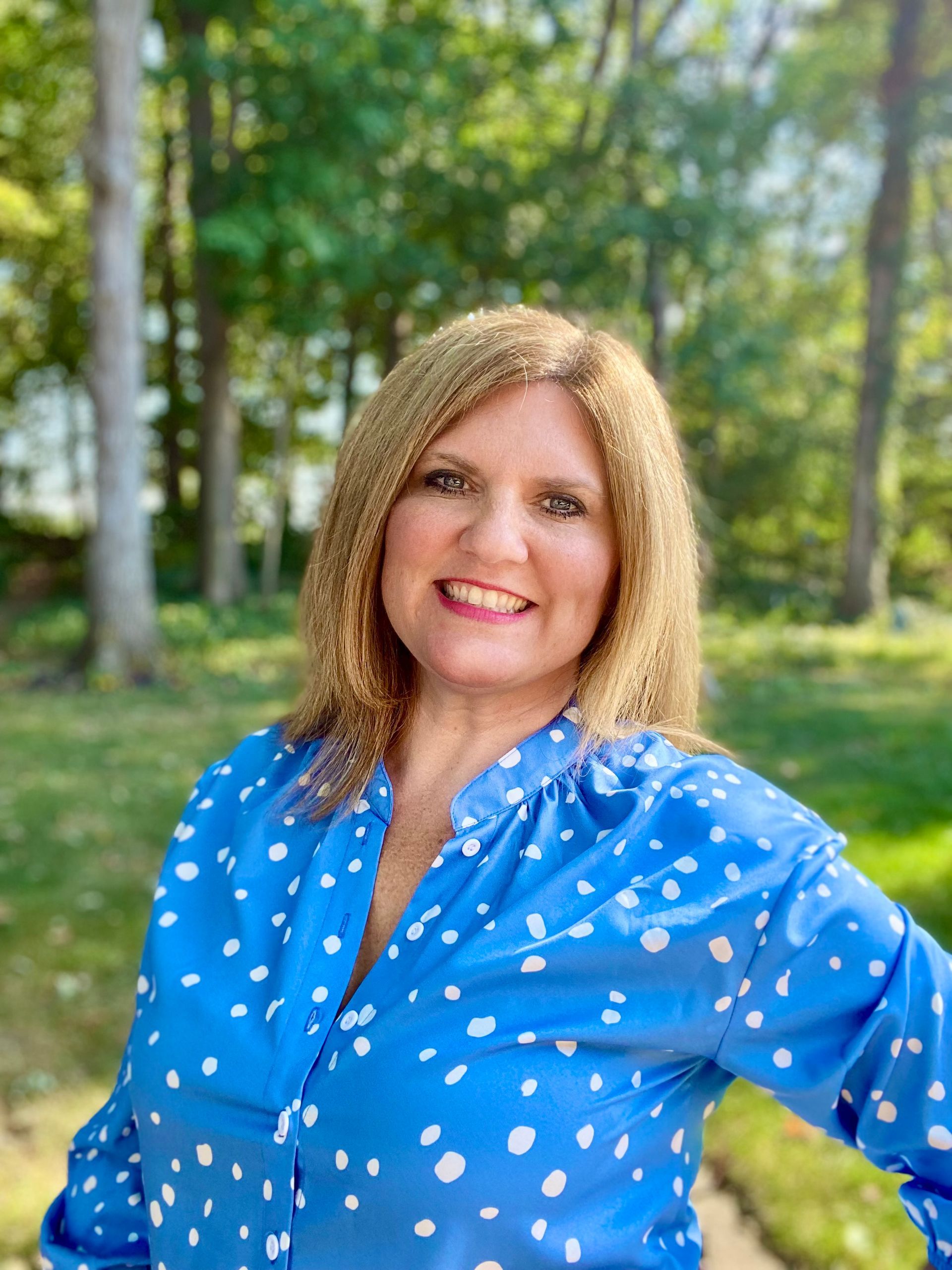 Woman with blonde hair smiling, wearing a blue polka-dot shirt, set in a green, outdoor setting.