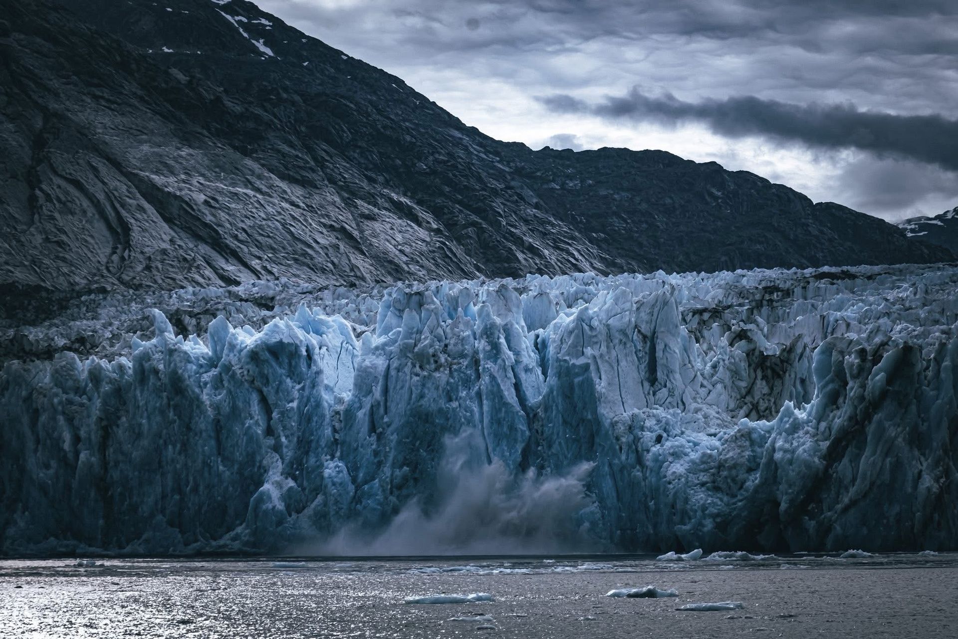 Glacier calving into the ocean, against a backdrop of dark mountains and a cloudy sky.