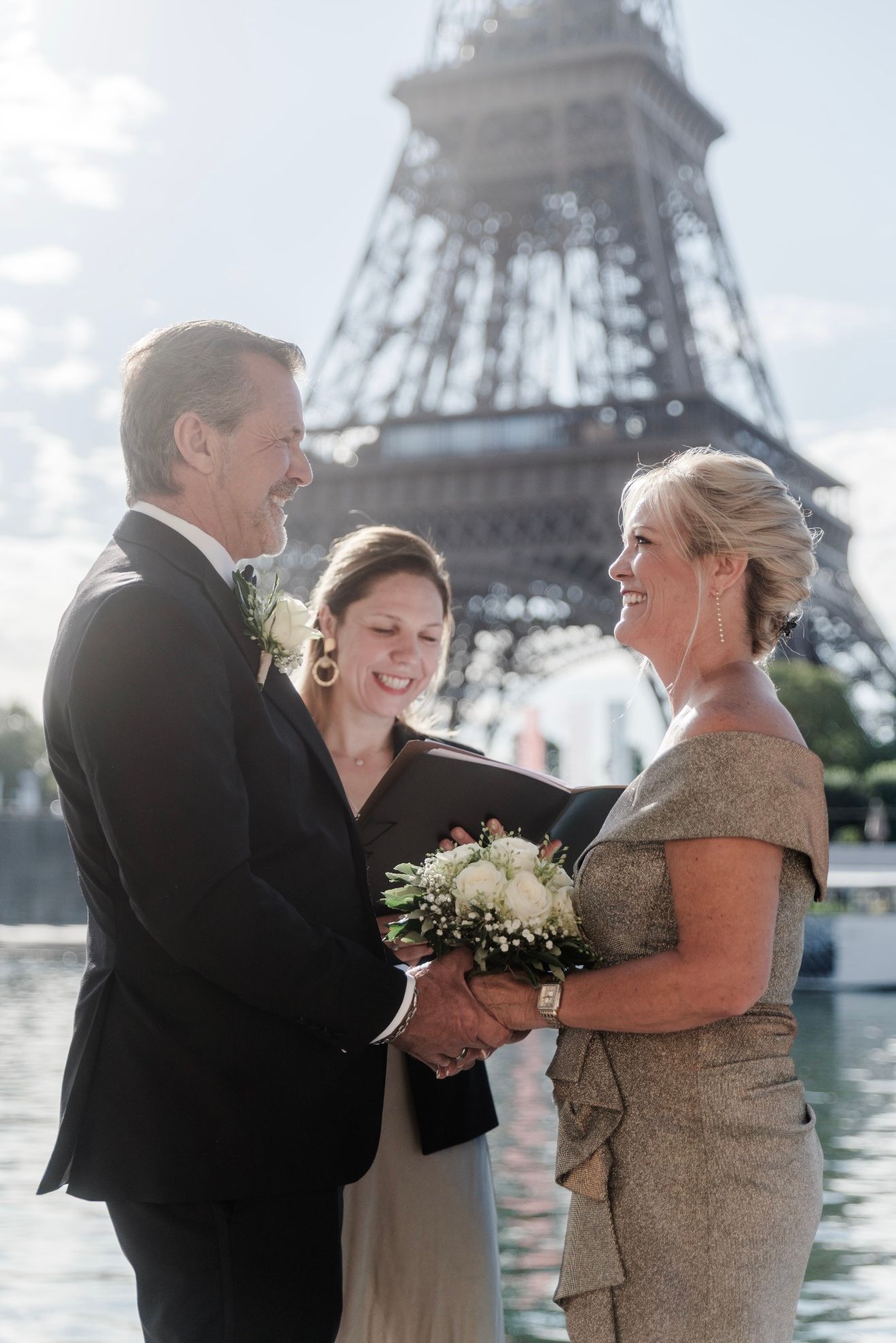 Couple at a wedding ceremony in front of the Eiffel Tower, holding hands. Officiant present, smiling.