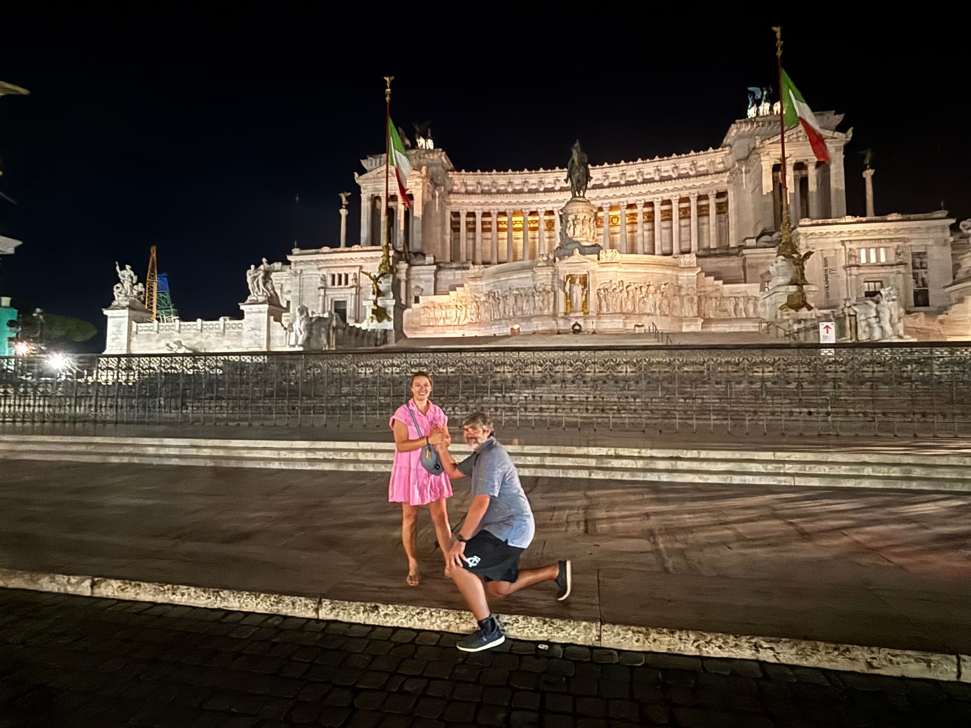 Man kneeling, proposing to woman in front of the illuminated Altare della Patria in Rome.