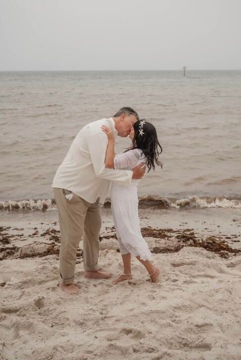 Couple kissing on a beach. Man in beige pants and white shirt, woman in white dress, ocean in background.