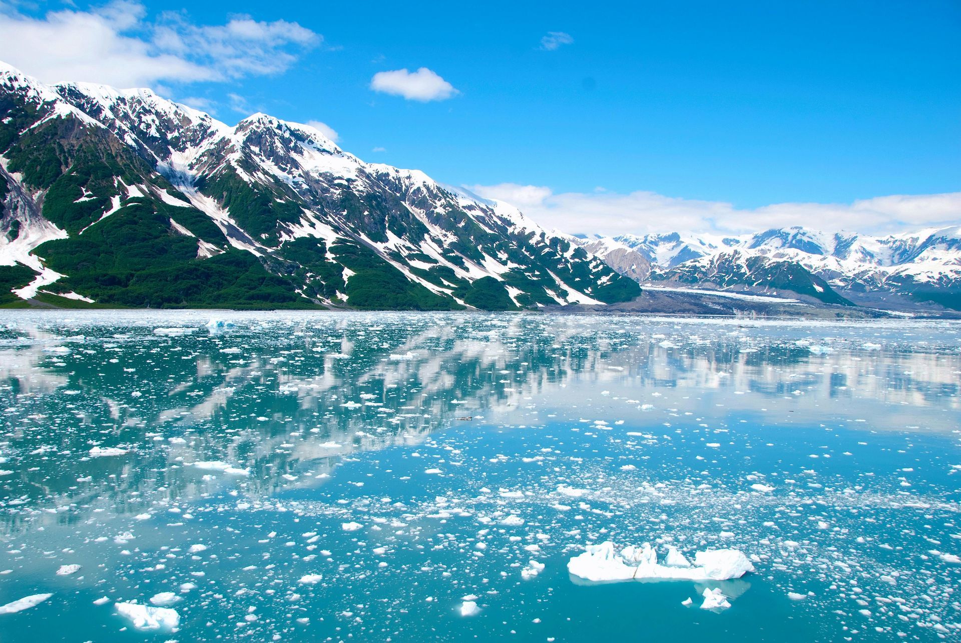 Snow-capped mountains reflected in icy blue water, with floating ice chunks under a clear sky.