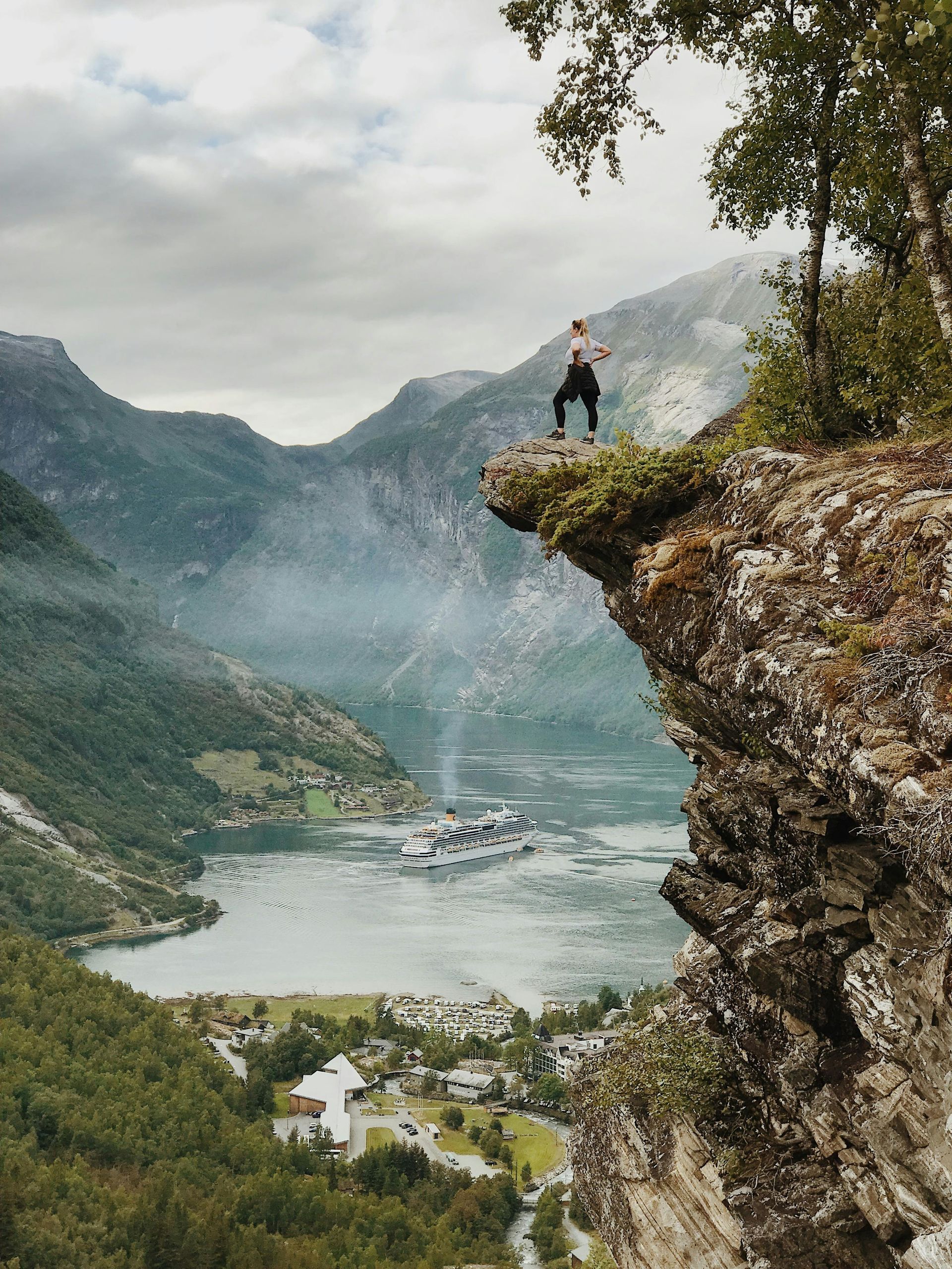 Person stands on cliff edge overlooking fjord and town.