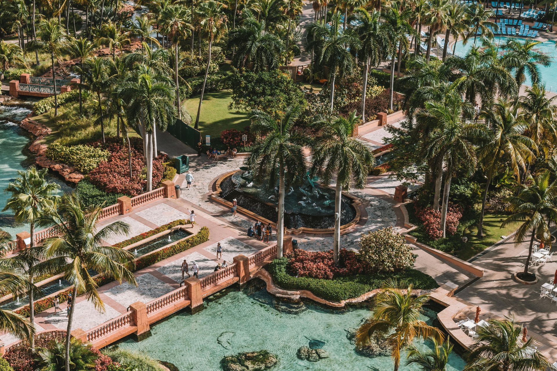 Aerial view of a tropical resort with pools, palm trees, pathways, and buildings under a bright sky.