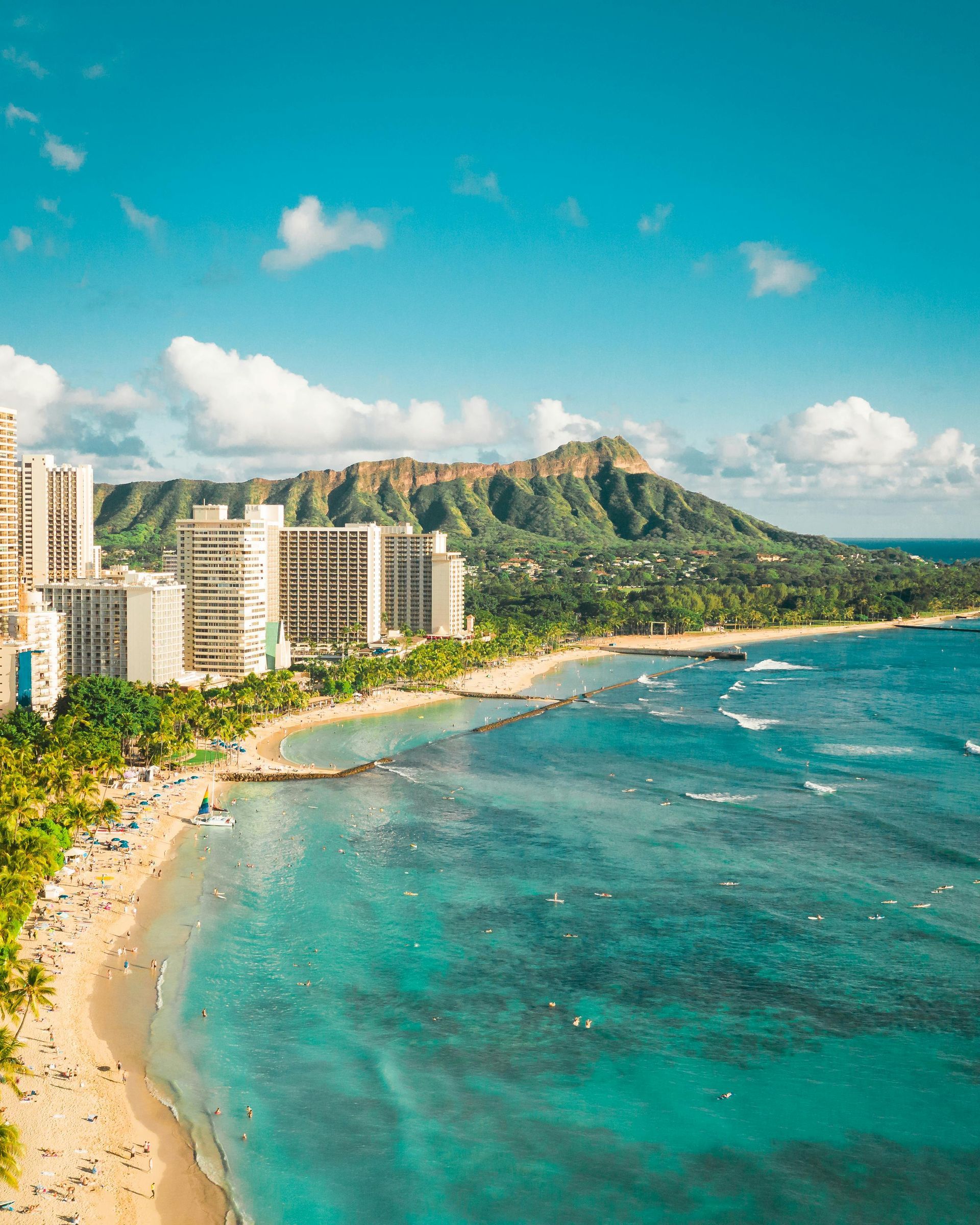 Waikiki Beach with turquoise water, white sand, and buildings, Diamond Head mountain in the background under a blue sky.