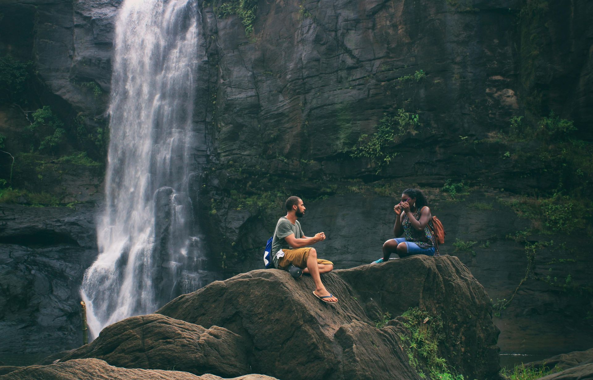 Two people sit on rocks near a waterfall. One holds something up, the other appears to look at it. Lush green backdrop.