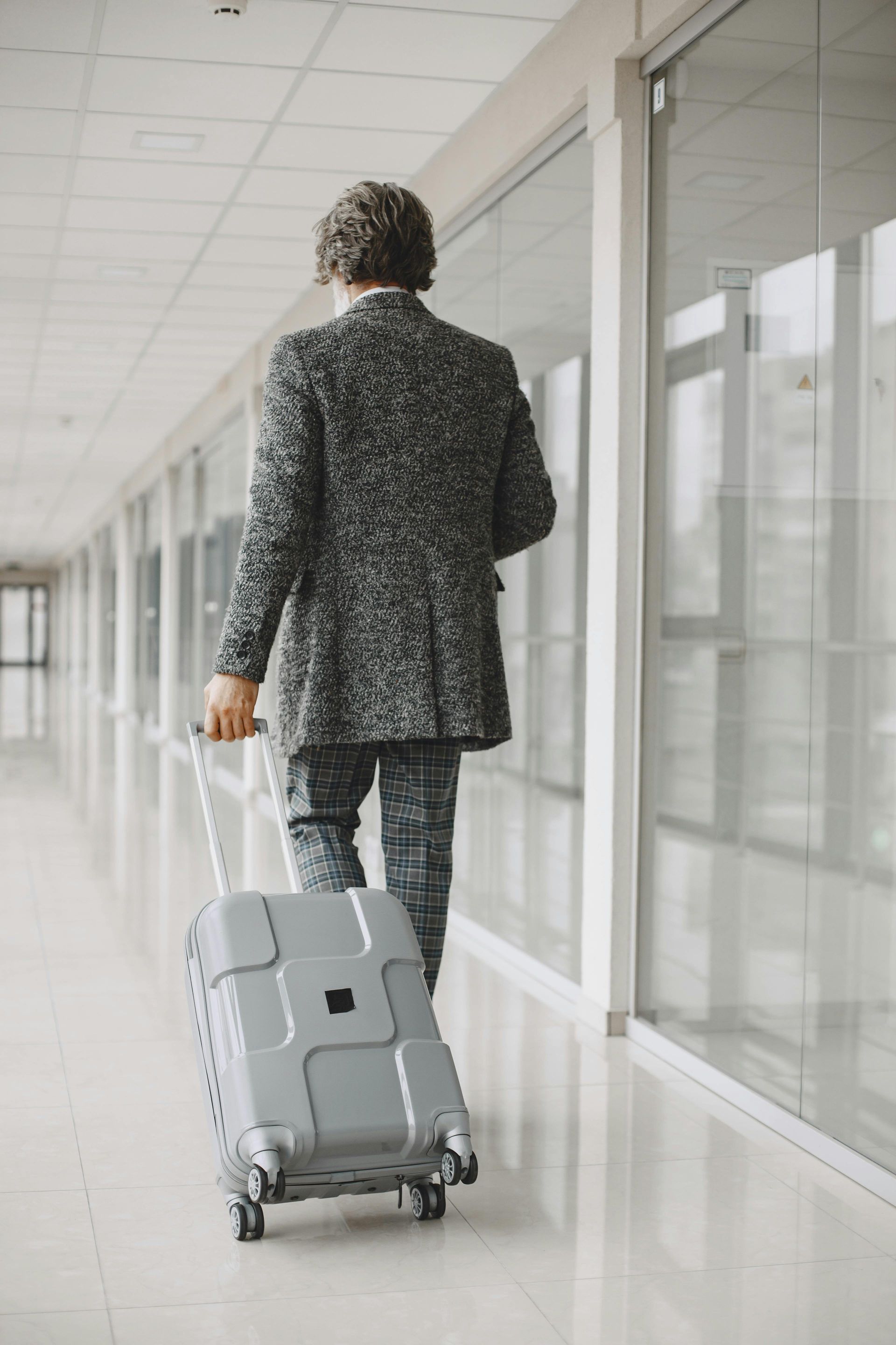 Person in patterned suit walking with rolling suitcase in a bright hallway.