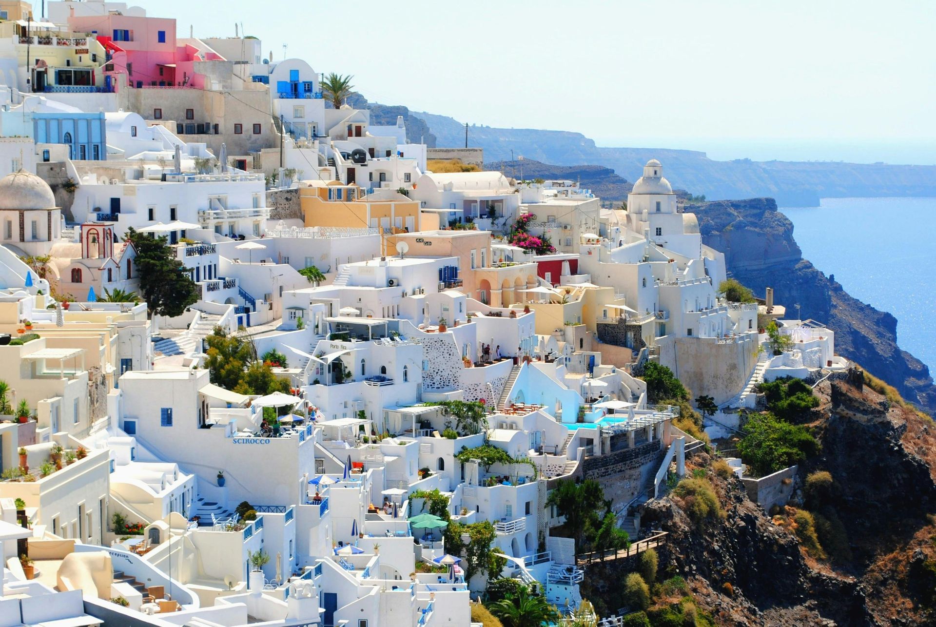 Whitewashed buildings with blue accents cascade down a cliffside, overlooking the Aegean Sea in Santorini.
