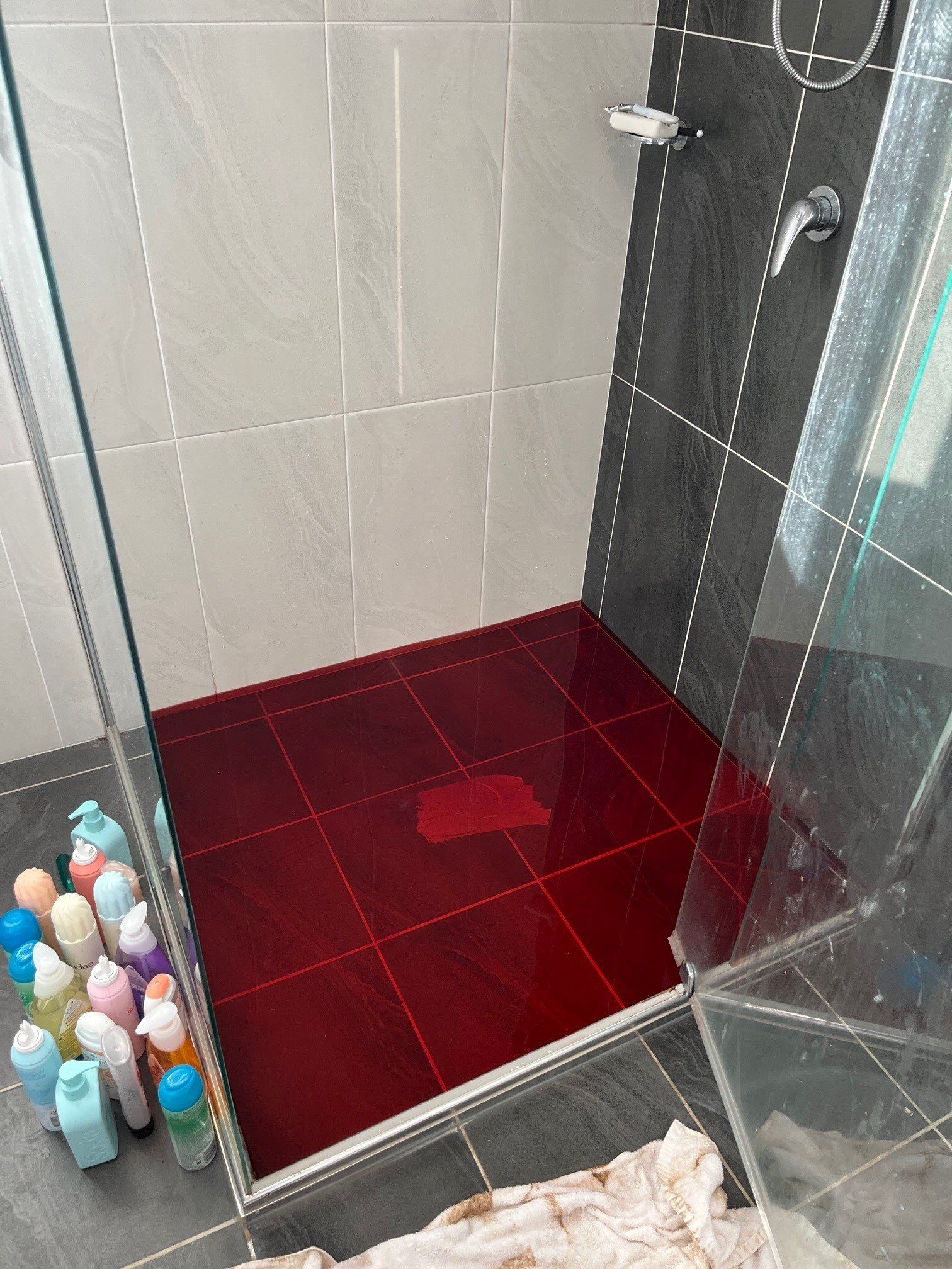 A shower floor filled with a deep red liquid, with a group of colorful toiletry bottles lined up on the bathroom floor. — Distinct Plumbing Services In Dapto, NSW