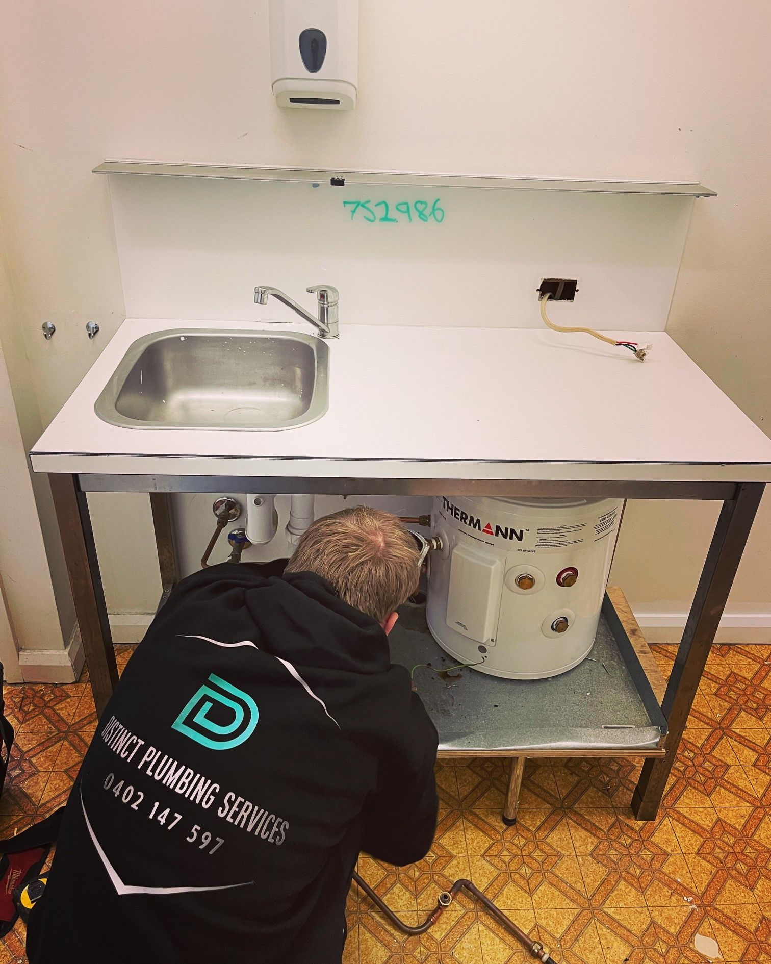 A plumber working under a sink cabinet beside a small electric water heater in a utility room. — Distinct Plumbing Services In Kiama, NSW