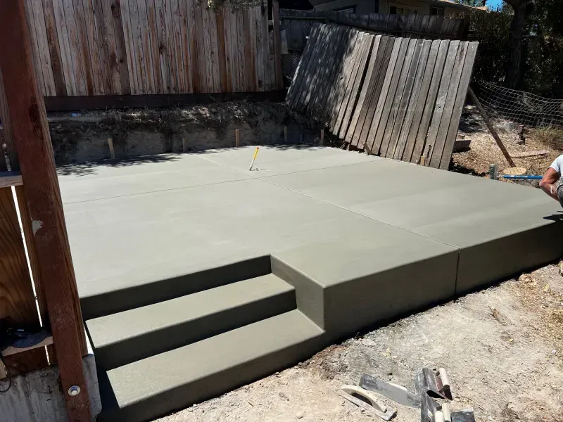Freshly poured concrete patio with three steps, near a wooden fence.