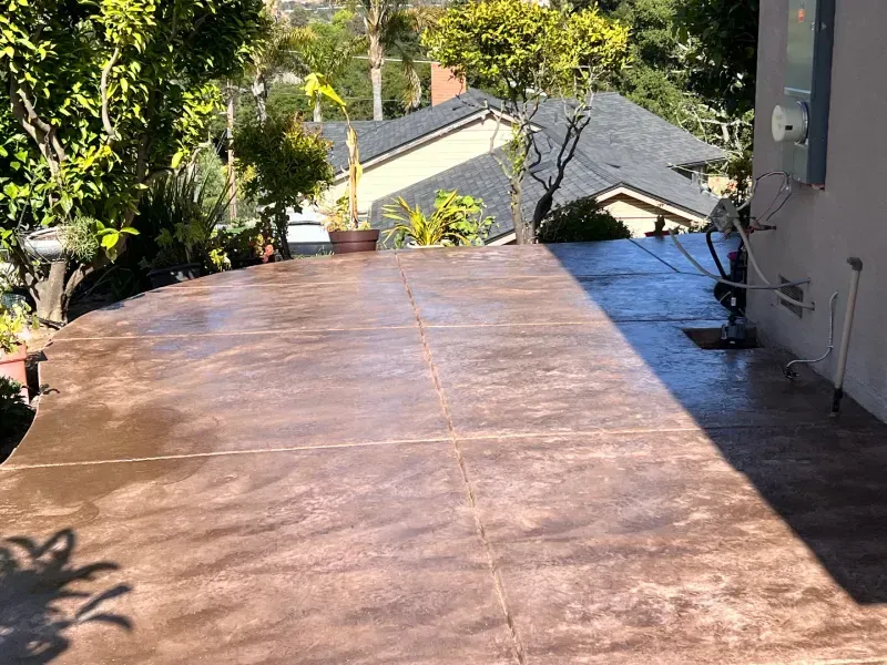 Brown stamped concrete patio with trees, house, and utility box.