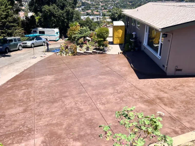 Brown-colored stamped concrete driveway and patio next to a house with a hillside view, a person standing.