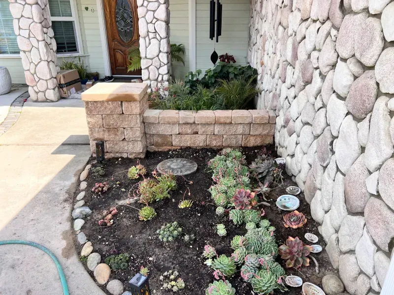 Succulent garden bed with stone wall and a tan brick planter near a house.