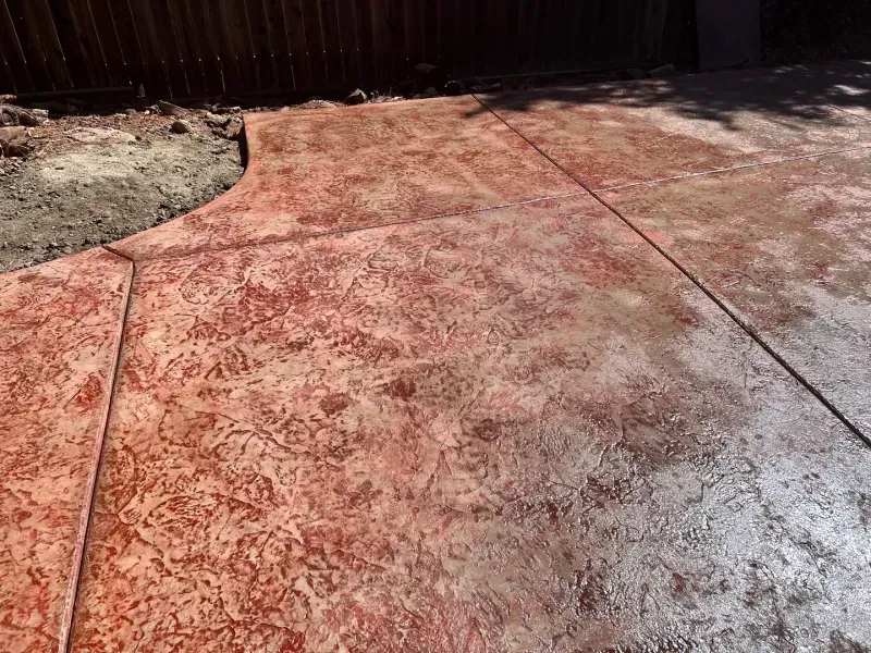 Stamped concrete patio with red and brown hues, near a fence and dirt.