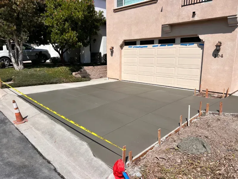 Freshly poured concrete driveway in front of a house. Yellow caution tape and construction stakes are present.